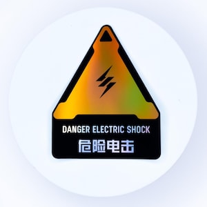 May include: A triangular holographic sticker featuring a black lightning bolt icon centered within a gold-toned triangle. Below the triangle, the text DANGER ELECTRIC SHOCK and the Chinese characters 危险电击 are printed in white on a black background.