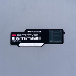 May include: A black rectangular label with rounded corners featuring the white text PROTOTYPE, SUPER-CAPACITOR, and IA DS4-1774822-5. It includes a red rectangle, two barcodes, small safety icons, and a white field with the serial number S/N 48-1508F.