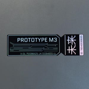 May include: A rectangular black sticker with a holographic finish featuring the white text PROTOTYPE M3, a circuit board graphic, the alphanumeric code X-OL-751286425-JK-51264855-IU, a pixelated symbol, and the vertical text ID: 78542030695.