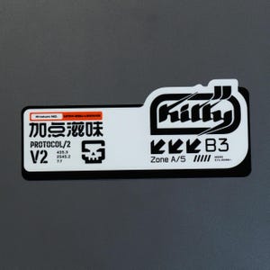 Puede incluir: Una pegatina rectangular blanca con texto y gráficos en negro. Incluye el texto "Killy", "PROTOCOL/2", "V2", "Zone A/5", "B3" y caracteres chinos. Un recuadro naranja contiene "Product NO. UF54-88U-L83W48". Se muestra un icono de una calavera.