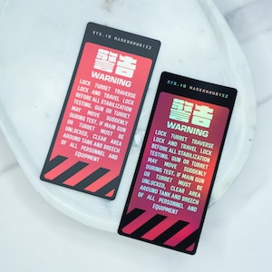 May include: Two rectangular warning labels with red and black diagonal stripes. The labels have the text "WARNING" in English and Japanese characters. The text warns about locking the turret and clearing the area during testing.