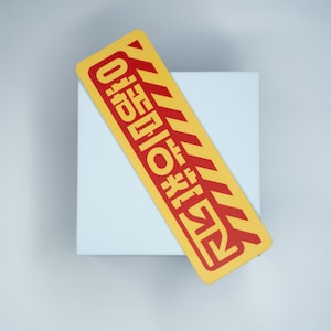 Puede incluir: Una pegatina rectangular amarilla con texto rojo y rayas diagonales rojas. La pegatina está colocada sobre un cuadrado blanco. El texto parece estar en un idioma extranjero. La pegatina tiene un diseño de advertencia o precaución.