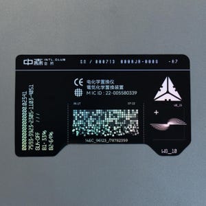 Puede incluir: Una tarjeta de identificación rectangular negra con esquinas redondeadas que presenta texto y gráficos en blanco. Muestra números de serie, el texto "IN UT", "07-22", "MIC ID : 22-005580339" y una cuadrícula central de pequeños cuadrados. Un logotipo triangular y un gráfico de ondas circulares están a la derecha.
