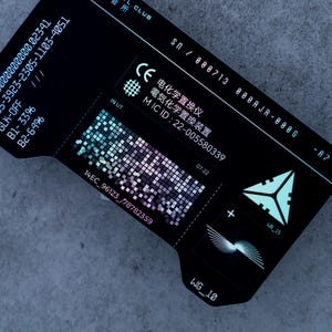 May include: A black electronic device with white text and a QR code. The device has a series of numbers and letters, including "MIC ID: 22-005580339". The device also has a geometric design and a plus sign.