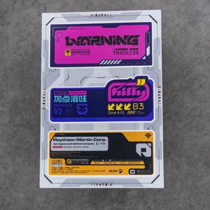 Puede incluir: Una hoja de pegatinas rectangular con tres etiquetas gráficas distintas. La etiqueta superior es rosa con el texto WARNING y TN45X139. La etiqueta central es azul con el texto Killy, Protocol/2 y Zone A/5. La etiqueta inferior es amarilla y negra con el texto Raytheon Martin Corp.