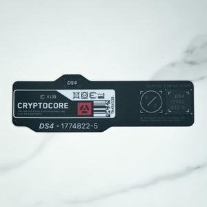 Puede incluir: Una etiqueta rectangular negra con texto y gráficos en blanco. Presenta el texto CRYPTOCORE, DS4 y 1774822-5. La etiqueta incluye un icono cuadrado rojo, un código de barras, símbolos técnicos y el texto SUPER-CAPACITOR.