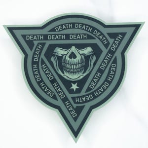 May include: A triangular patch in shades of sage green and dark charcoal grey. The design features a central human skull graphic above a small star, surrounded by a repeating border of the word DEATH printed in a bold, sans-serif typeface.