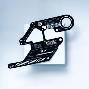 Może przedstawiać: Czarny, wykrawany element z napisem "NANOSWITCH" i szczegółami technicznymi. Element ma unikalny kształt z okrągłym otworem i różnymi wycięciami. Przedmiot umieszczony na białej powierzchni.