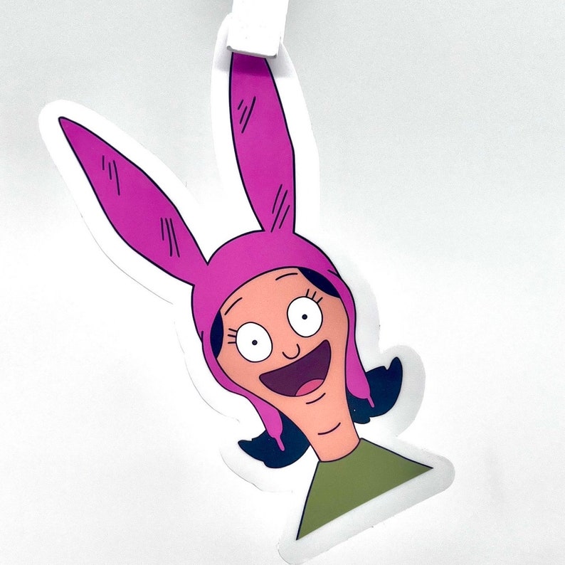 May include: A sticker of a cartoon character with pink bunny ears, a pink hat, and a big smile. The character has brown hair, brown eyes, and is wearing a green shirt.