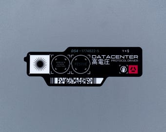 May include: A black rectangular sticker with white text and graphics. It features a sunburst icon, circular target markings, a barcode, and the text DS4-1774822-5, DATACENTER, PROTOCOL DRIVER, and Japanese characters for high voltage.