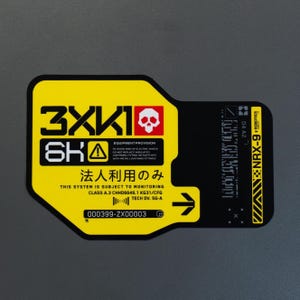 Peut inclure: Un autocollant rectangulaire aux coins arrondis avec un design jaune et noir. Il affiche le texte 3XK1, une icône de crâne, des caractères japonais et des spécifications techniques incluant NRX-9 ROBOTICS et le numéro de série 000399-ZX00003.