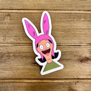 May include: A sticker featuring a cartoon character with pink bunny ears, a green shirt, and a wide smile.