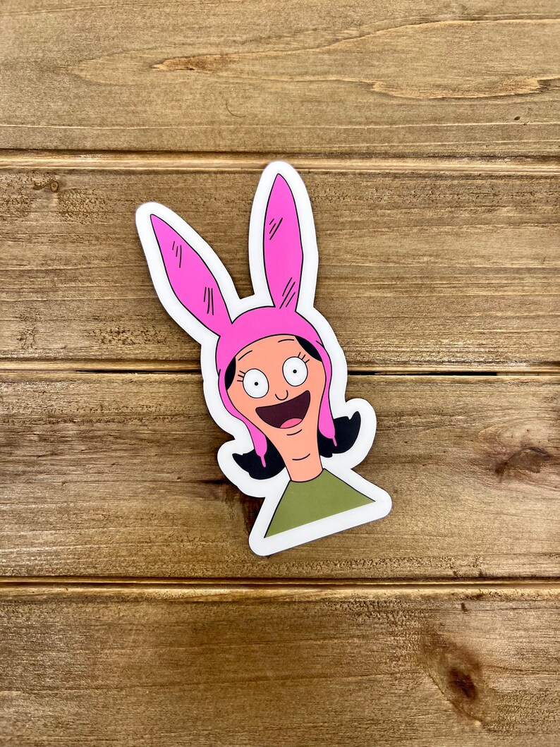 May include: A sticker featuring a cartoon character with pink bunny ears, a green shirt, and a wide smile.