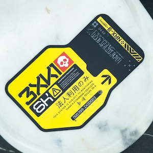 May include: A black and yellow sticker with the text "3XK10 6KA" and other technical details. The sticker features a skull icon, an arrow, and the text "NRX-9". The sticker is on a white marble surface.