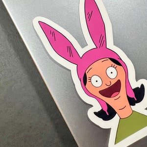 May include: A sticker featuring a cartoon character with pink bunny ears and a big smile. The character has brown hair and is wearing a green shirt.