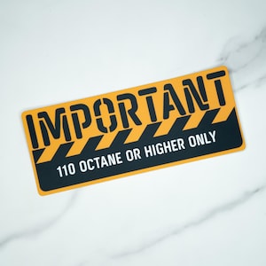以下が含まれることがあります： 「重要」の文字が大きく黒いブロック体で書かれた、長方形の黄色と黒のサイン。その下に「110オクタン以上のみ」と書かれています。サインには黒と黄色の斜めストライプが入っています。