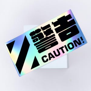 May include: A holographic sticker with black text and design elements. The sticker reads "CAUTION!" in bold, capitalized letters. Above the word "CAUTION!" is a series of black, stylized characters. Two black diagonal stripes and a triangle are on the left side.