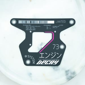 May include: A black, flat, T-shaped technical decal featuring white text, a barcode, and a pink accent line. The text includes 'LIMITED', 'TECHNICAL SPEC', 'ITEM NUMBERS', 'SYSTEM', '73', 'エンジン', '12 SEPT 2089 DRIVER VERSION S57TVM_33', and 'DECAY'.