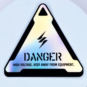 May include: A triangular holographic sticker with a black border and a black lightning bolt icon. The text DANGER is printed in black, followed by the text HIGH VOLTAGE. KEEP AWAY FROM EQUIPMENT. The sticker has a reflective, iridescent finish.