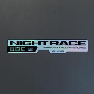 May include: A rectangular holographic sticker with a dark background featuring the text NIGHT RACE in bold capital letters. The sticker includes the text WARRANTY VOID IF REMOVED, the code MT21 - XXB52, and small technical icons on the left side.