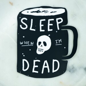 May include: A black sticker shaped like a coffee mug with a white skull graphic in the center. The text reads SLEEP WHEN I'M DEAD in white capital letters. The design features small white dots scattered across the black surface of the mug.