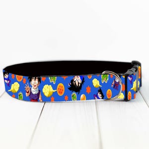 May include: Blue dog collar featuring colorful cartoon characters and symbols from the Dragon Ball Z anime series. The collar has a black interior and a silver metal D-ring for leash attachment. The collar is made of durable material.