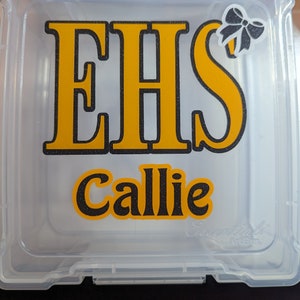 May include: A clear plastic storage box with a yellow and black EHS logo and the name Callie written below. A small black bow is in the top right corner.