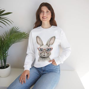 Può includere: Felpa bianca con una grafica di un coniglio che indossa occhiali a forma di cuore. Il coniglio è grigio con orecchie e naso rosa.