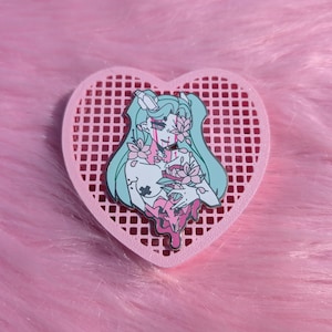 May include: A pink heart-shaped pin holder with a metal pin featuring a teal-haired anime-style character with pink floral accents and dripping pink details. The pin is set against a pink, fluffy background.
