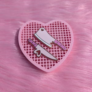 May include: Two pink enamel pins in the shape of a cleaver and a knife, with heart-shaped cutouts, rest on a pink heart-shaped grid. The pins have gold accents and are set against a fluffy pink background.