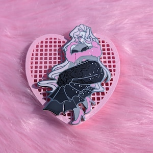 May include: A pin featuring a stylized illustration of a woman in a black dress with rose details, a pink fur stole, and pink heels. The pin is set on a pink heart-shaped background.
