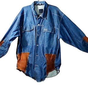 Può includere: Giacca di jeans blu con toppe in pelle scamosciata marrone sui gomiti e sulla parte anteriore inferiore. La giacca ha una chiusura con bottoni, due tasche sul petto e le parole "Jean John" ricamate sul petto.