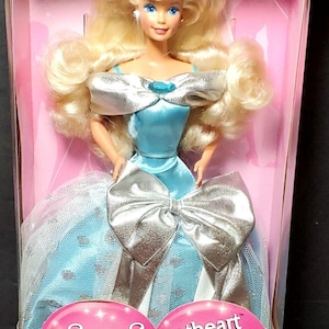 Puede incluir: Una muñeca Barbie Silver Sweetheart en una caja rosa. La muñeca tiene cabello rubio, ojos azules y viste un vestido azul claro con detalles plateados. La caja tiene el texto "Exclusive Limited Edition SEARS" y "Silver Sweetheart Beauté Argentée Barbie".