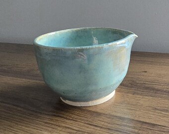 May include: A handmade ceramic bowl with a turquoise glaze. The bowl has a rounded shape with a spout and a white base. The bowl is sitting on a wooden surface.