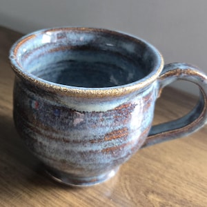 May include: A handcrafted ceramic mug with a rounded body and a handle. The mug has a blue and brown glaze with a gold rim. The interior is a darker blue. This stoneware mug is ideal for coffee or tea.