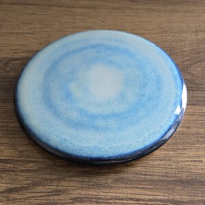 Peut inclure: Un dessous de verre rond en céramique avec un motif aquarelle bleu et blanc. Le dessous de verre a un bord bleu foncé et une finition brillante. Le motif présente des cercles concentriques bleus clairs et foncés, créant un effet tourbillonnant.