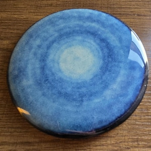 May include: A round, ceramic coaster with a blue and white watercolor design. The coaster has a dark blue rim and a glossy finish. The center of the coaster is a lighter shade of blue, creating a gradient effect.