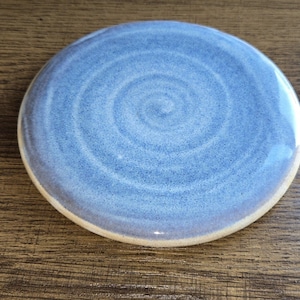 May include: A round, ceramic coaster with a light blue, spiral design. The coaster has a speckled texture and a cream-coloured rim. The coaster is on a wooden surface.