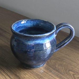 Peut inclure: Une tasse en céramique faite à la main avec un glaçage bleu foncé. La tasse a un corps arrondi, une anse robuste et une finition brillante. La couleur passe d'un bleu marine foncé à un bleu plus clair et plus vif près du bord. Parfait pour le café ou le thé.