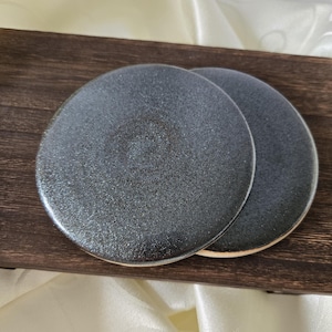 May include: Two round, dark grey coasters with a speckled texture are displayed on a dark brown wooden tray. The coasters have a metallic sheen and a light-coloured edge. The background is a white, textured fabric.