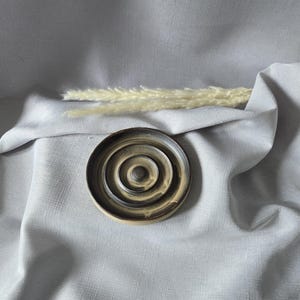 May include: A handmade ceramic soap dish with a concentric circle design in shades of brown and beige. The dish is placed on a grey fabric background, with a sprig of dried ornamental grass. The dish is approximately 10 cm in diameter.