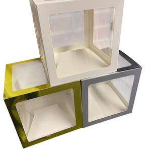 Könnte beinhalten: Ein Stapel von vier quadratischen Backboxen mit transparenten Fensterpaneelen. Die Boxen sind in Weiß, Gold, Grau und Dunkelgrau gehalten. Die leeren Boxen sind gebrauchsfertig.