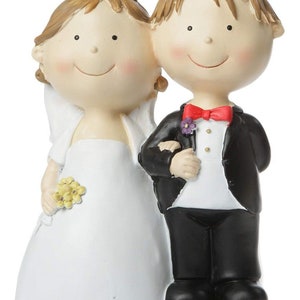 Könnte beinhalten: Ein weißer Hochzeitstortenaufsatz mit einem Cartoon-Brautpaar. Die Braut trägt ein weißes Kleid und eine Blumenkrone. Der Bräutigam trägt einen schwarzen Smoking mit einer roten Fliege.