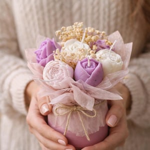 Può includere: Una composizione di candele decorative con diversi fiori in cera nelle tonalità lavanda, rosa pallido e bianco, arricchita da fiori secchi. I fiori sono inseriti in un contenitore rosa avvolto in carta traslucida e legato con dello spago.