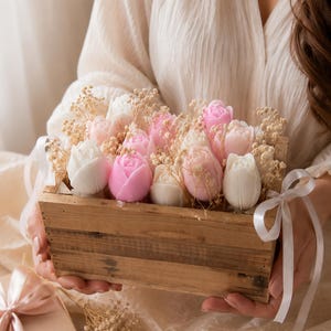 Könnte beinhalten: Eine Holzkiste gefüllt mit Seifenrosen in Rosa- und Weißtönen, akzentuiert mit getrocknetem Schleierkraut. Die Kiste ist mit einem weißen Band gebunden. Die Anordnung wird von einer Person gehalten.