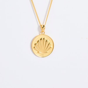 Op de afbeelding: Een goudkleurige ketting met een ronde hanger met een verhoogd, reliëf schelpontwerp in het midden. De hanger hangt aan een fijne, dunne goudkleurige ketting die via een klein, eenvoudig oogje aan de bovenkant van de hanger is bevestigd.