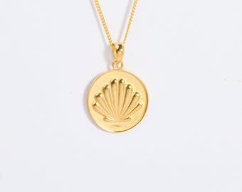May include: A gold-toned necklace featuring a circular pendant with a raised, embossed seashell design in the center. The pendant hangs from a delicate, thin gold-toned chain that connects to the top of the round charm via a small, simple bail.
