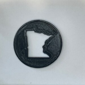 Può includere: Un sottobicchiere nero stampato in 3D con il contorno dello stato del Minnesota.