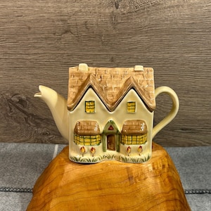 Peut inclure: Une théière en céramique en forme de cottage avec un toit brun, des murs blancs et des fenêtres jaunes. La théière est posée sur une base en bois.