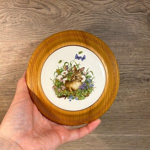 May include: A round wooden plaque with a white ceramic tile inset. The tile features a hand-painted illustration of a brown rabbit sitting in a field of green grass and colorful flowers. There are two blue butterflies flying above the rabbit.